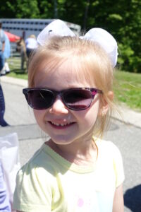 A young girl with blonde hair smiles brightly while wearing purple sunglasses and a white bow in her hair. She is outdoors on a sunny day, dressed in a light yellow shirt, with a background of greenery and people walking by.