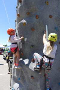 Two children wearing safety helmets and harnesses climb a tall outdoor rock climbing wall. One child is wearing a pink helmet and the other a yellow helmet. They are mid-climb, each using colorful holds on the grey textured wall. A vehicle and sky are visible in the background.