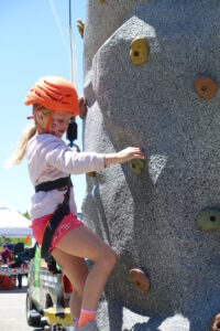 A young girl wearing a helmet and harness climbs an outdoor rock wall. She grasps colorful handholds while looking focused. She’s dressed in a long-sleeve shirt and pink shorts. Sunlight and a clear blue sky indicate it's a sunny day.