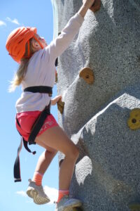A young girl wearing a helmet and safety harness climbs a rock wall on a sunny day. She is wearing a white sweatshirt, red shorts, and orange socks, with her blonde hair tied back. The sky is clear blue with a few white clouds in the background.