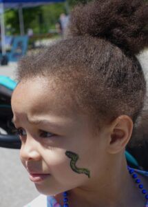 A young child with curly hair and a high bun looks to the side while outdoors. They have a green snake painted on their cheek and wear blue beads around their neck. The background features a tent and bright daylight.