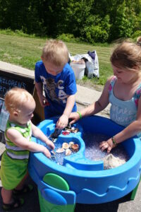Three children are playing with a sensory table filled with various small objects, including shells and beads. The table is blue and has multiple compartments. Behind them, a grassy area and some trees are visible. A sign reading "children's" is partially visible.