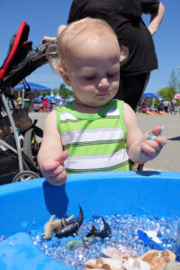 A toddler in a green and white striped tank top is playing with a sensory bin filled with water beads and toy sea creatures. A stroller is visible in the background, along with people and tents, indicating an outdoor event or fair. The child is focused on the toys.
