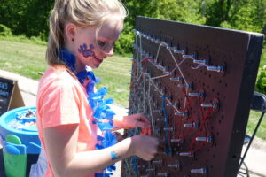 A girl with face paint and a blue lei stands outdoors, manipulating a large vertical pegboard with elastic cords. Nearby, a blue container holds various beads or trinkets. The scene suggests a playful and interactive activity on a sunny day.