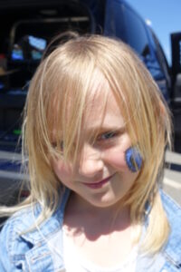 A young child with blonde hair is standing outdoors. They are wearing a blue denim jacket over a white shirt. The child has a blue and yellow face paint design on their cheek and is smiling slightly. The background shows the back of a vehicle and some out-of-focus scenery.