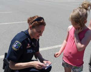 A police officer kneels on the ground, writing on a notepad while a young girl in a pink tank top and patterned shorts watches. They are outdoors in a parking lot on a sunny day. The girl has her hand near her face, and the officer is wearing a uniform with a badge.