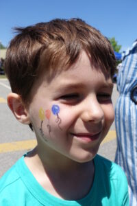 A young boy with short brown hair is smiling outdoors. He has a face painting of three colorful balloons on his cheek. The background shows a blue sky and indistinct figures. He is wearing a turquoise shirt.