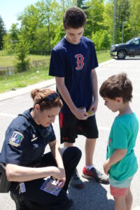 A female police officer kneels and shows papers to two boys in a parking lot. The older boy, wearing a navy t-shirt and red sports shorts, and the younger boy, in a green t-shirt, look at the papers. Trees and a parked car are visible in the background.