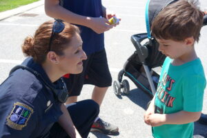 A female police officer kneels to talk with a young boy wearing a green shirt in a parking lot. The boy is facing the officer, who is smiling and holding a conversation. Nearby, another child, partially visible, stands holding a toy, and a stroller is seen in the background.