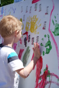 A young child with blond hair and a bandage on their arm is pressing their hand onto a large piece of white paper covered with colorful handprints and paint streaks. The child is wearing a white t-shirt with blue and black stripes on the sleeves.