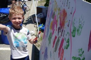 A young child with curly hair, wearing a white t-shirt, smiles and holds a paintbrush while standing next to a large board filled with colorful handprints and brush strokes. The scene appears to be an outdoor activity or fair.