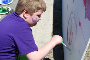 A child with short blond hair and a purple shirt is painting on a large canvas outside. He is using a green-handled brush to apply paint. There is a colorful face paint design on his cheek and paint supplies are visible on the ground beside him.