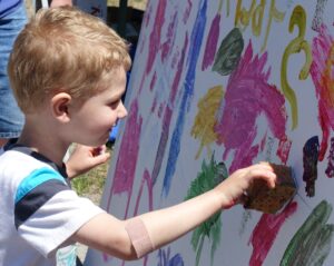 A young child with a band-aid on their arm is using a sponge to paint on a large canvas filled with colorful brushstrokes and splashes of paint. The child appears focused and engaged in the activity. The scene suggests an outdoor art activity.