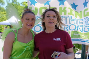 Two women are standing outdoors in front of a banner. One is dressed in a Tinkerbell costume, wearing a green dress and light green fairy wings, and the other is in a maroon t-shirt holding a phone. They are both smiling. The banner behind them has blue stars and text.
