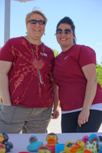 Two smiling people in maroon shirts pose together at a booth with colorful rubber ducks in front of them. They are outdoors on a sunny day, and both wear sunglasses. The person on the left has short blond hair and the person on the right has long dark hair tied back.