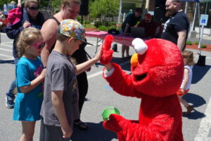A person dressed as Elmo high-fives a young boy wearing a floral baseball cap. A young girl in pink sunglasses stands beside him, smiling. Other people, including a bearded man in the background, are also present at this outdoor event.