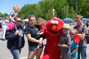 A group of people, including children, are posing outdoors with a person dressed in a red character costume that resembles Elmo from "Sesame Street." Some children are smiling, and one child is being carried on a woman's shoulders. The weather appears sunny, and trees are visible in the background.