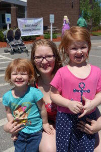 A woman with glasses and a red top crouches and smiles between two young girls. The girl on the left wears a teal shirt and shorts, while the girl on the right wears a pink shirt and blue polka-dotted skirt. They are outside at an event with a sign in the background.