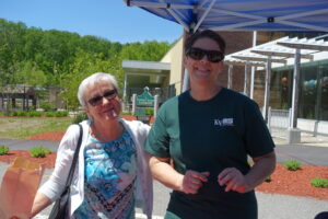 Two women stand outdoors under a canopy. One woman, on the left, has short white hair, sunglasses, and a blue floral shirt, holding a paper bag. The other, to the right, has dark hair in a bun, wears sunglasses, and a dark green shirt with a logo, smiling. Trees and a building are visible in the background.