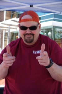 A man wearing a backwards orange cap, sunglasses, and a maroon T-shirt with a "KVCAP" logo points both index fingers toward the camera. He is smiling and standing under a tent, possibly at an outdoor event.