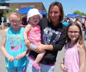 A woman is standing outside with three children. The woman is holding a toddler wearing a white hat and pink outfit. Two older children, both in glasses and pink tank tops, stand beside them. All are smiling. In the background, there is an Educare building.