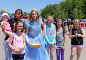 A group of children and one adult pose outdoors at an event. A girl in a blue princess dress stands in the center with a basket. Sunny sky and greenery suggest it's a warm day. Two children wear glasses, and one adult holds a baby in the back.