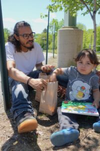 A man with glasses and long hair sits on the ground near a tree, holding a brown paper bag. A young child with a book sits beside him, wearing a gray shirt with a colorful character on it, blue pants, and blue shoes. They appear to be enjoying a casual outdoor moment.