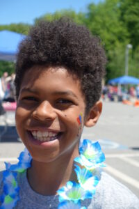 Smiling child wearing a blue and green lei and face paint with a colorful rainbow design, standing outdoors with a blurred background of trees, tents, and other people.