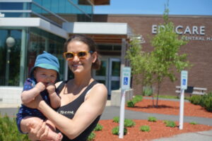 A woman with sunglasses holds a baby wearing a blue hat in front of a building with a sign that reads "EDUCARE" in the background. The scene is outside on a sunny day with clear skies and some greenery visible around the area.