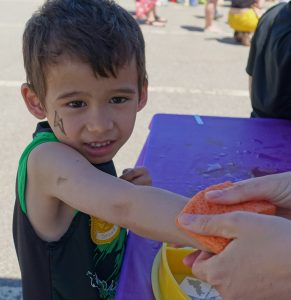 A young boy with face paint is having his arm washed with a sponge by an adult at an outdoor event. He is seated at a purple table, and the background shows other people and activities.
