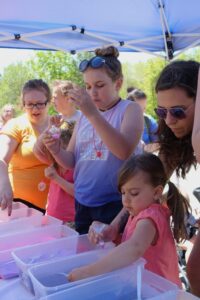 A group of children and adults engage in a craft activity under a blue canopy. Several plastic bins filled with colorful materials are on the table. The children are focused on their tasks, while adults assist and observe. A sunny day with green trees in the background.
