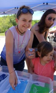 A young girl smiling while playing with colored sand at an outdoor event. She is wearing sunglasses on her head, a purple tank top, and a choker necklace. Another child and an adult stand nearby, engaged with the activity. Trees and a tent are visible in the background.