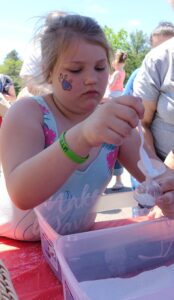 A young girl with a paw print painted on her cheek concentrates on scooping powdered substance into a container using a plastic spoon. She is wearing a colorful tank top and has a green wristband. Other people are blurred in the background on a sunny day.