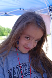 A young girl with long brown hair and face paint on her cheek is smiling mischievously. She is wearing a gray hoodie with text and a red lanyard, standing under a blue canopy with nature in the background.