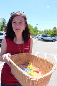 A young woman is standing in a parking lot on a sunny day, smiling and holding out a large wicker basket filled with colorful toys. She is wearing a red sleeveless shirt with the word "EDUCARE" printed on it. Sunglasses rest atop her head.