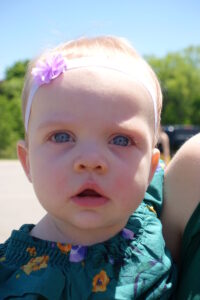 A close-up of a baby with blue eyes, wearing a light purple flower headband and a green floral dress. The baby appears to be outdoors on a sunny day with a blurred background of green foliage and a clear blue sky.