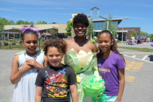 A person dressed as Tiana from "The Princess and the Frog" stands outdoors with three smiling children. The group appears happy and is in front of a building under a clear blue sky. The children are wearing casual clothes and look excited.