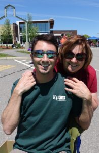 A man and woman smile at the camera in an outdoor setting. The man is wearing a green shirt with "KV CAP" on it and sunglasses, while the woman, also in sunglasses, stands behind him with her hands on his shoulders. A building and trees are visible in the background.