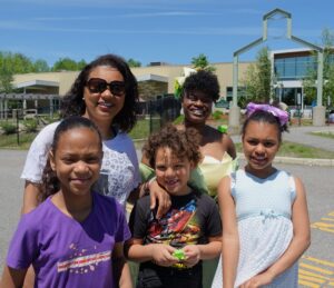 A group photo taken outside on a sunny day. Two women and three children are smiling in front of a building with a tall entrance arch. The woman on the left wears sunglasses and a white shirt, while the other wears a green dress. The children wear colorful casual clothes.