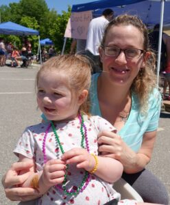 A woman with glasses and a child wearing a polka dot shirt sit together at an outdoor event on a sunny day. The child is adorned with colorful beads and is smiling. People and tents with activities, like "Sand Art," are visible in the background.