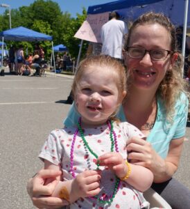 A woman and a young girl are smiling at an outdoor event. The girl is wearing a white polka-dot shirt, colorful beaded necklaces, and a yellow bracelet. The woman is wearing glasses and a striped shirt. There are booths and people in the background.