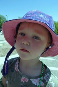 A young child with short hair wearing a sunhat and green beaded necklace looks at the camera. The child is outside on a sunny day, with a background of trees and pavement.