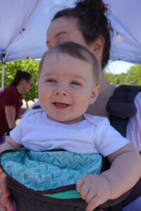 A baby with a happy expression, wearing a white shirt and sitting in a green and blue patterned carrier, with an adult holding them close under a blue tent. Another person is visible in the background. Trees and a sunny sky are seen in the distance.