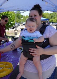 A woman with a tattoo on her left arm is holding a smiling baby in a front carrier under a tent at an outdoor event. Another person is seen in the background wearing a red shirt and sunglasses, setting up items on a purple table.