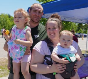 A smiling family of four poses outdoors on a sunny day. The mother is holding a baby in a front carrier, while the father carries a young girl who wears a floral romper and is holding a toy. They are standing in front of a blue canopy.