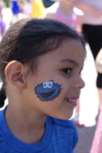 A young child with dark hair in braids is smiling and looking to the side. Their cheek is painted with a blue cookie monster face. The child is wearing a blue shirt and appears to be outside on a sunny day. Other children are visible in the background.