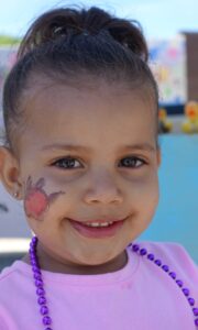 A young girl with a bun hairstyle smiles at the camera. She has a face painting of a red flower on her cheek and is wearing a pink shirt and purple beaded necklace. The background appears to be outdoors with colorful decorations in the distance.