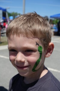 A young boy with short blond hair smiles outdoors. He has a green snake painted on his left cheek. The background shows a sunny day with blue sky, tents, and people blurred out.