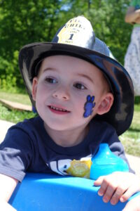 A young boy, with a badge sticker on his shirt and face paint on his cheek, wears a plastic firefighter helmet and smiles brightly while playing outside on a sunny day.