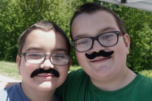 Two boys with short hair and glasses smile while wearing fake black mustaches. They are outdoors on a sunny day with green trees in the background.